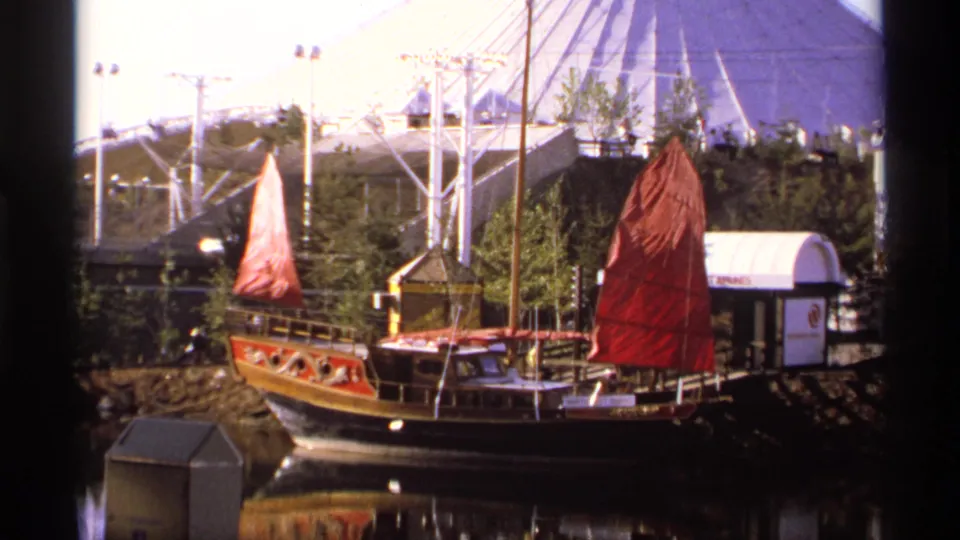 1974 8mm fairground view of a Chinese junk-style boat with red sails on water, backed by a geodesic dome and trees.