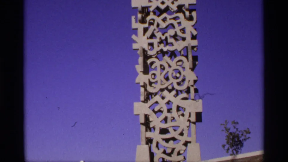 1970s 8mm close-up of a white geometric monument, centered against blue sky with a small tree visible near the base.