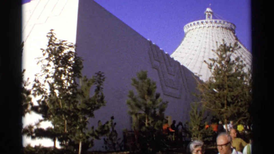 Wider 8mm view of modernist building walls, landscaped trees, visitors at ground level, and a ribbed dome behind them.