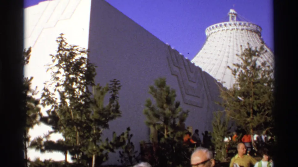 Wider frame showing visitors among trees below angular modern architecture, with a large ribbed dome in the background.