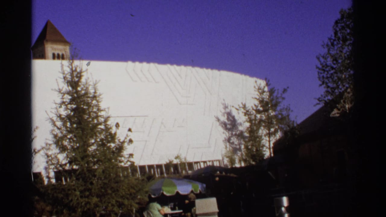 1974 8mm frame of a white modernist building in Germany, partly obscured by trees, with a small tower and umbrellas near the foreground.