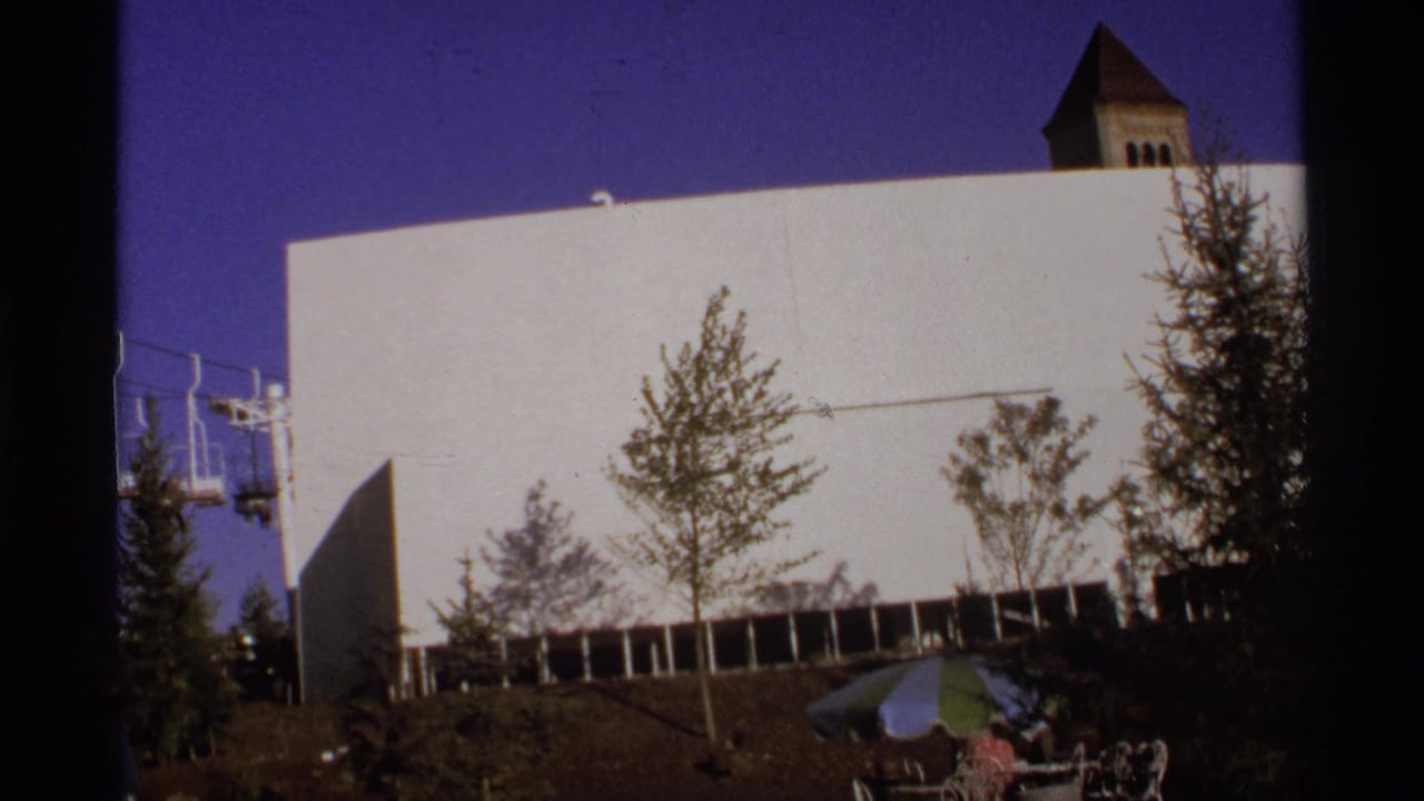 White building facade with trees, a small roof tower, umbrellas, and people near the lower edge.