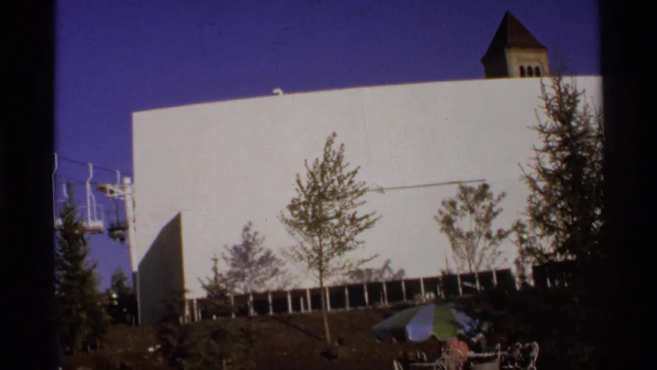 White modernist building in Germany seen from a visitor angle, with trees, a small roof tower, umbrellas, and small visible figures below.