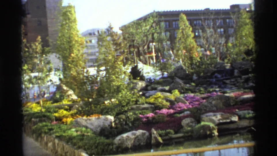 1974 8mm view of a landscaped garden in Germany with bright flowerbeds, rocks, trees, water, and urban buildings beyond.