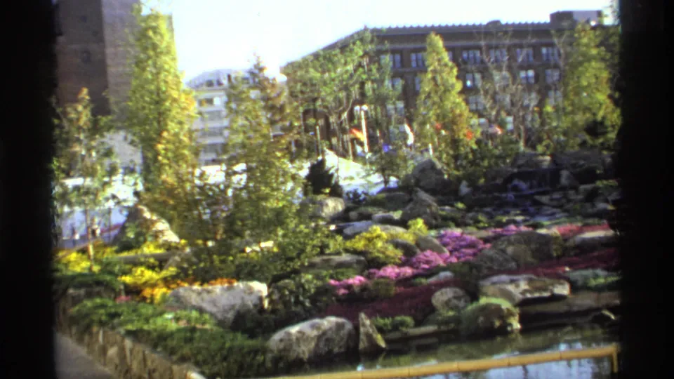 Visitor-shot 8mm frame of a colorful German garden, showing flowerbeds, rocks, water, trees, and urban buildings in the background.