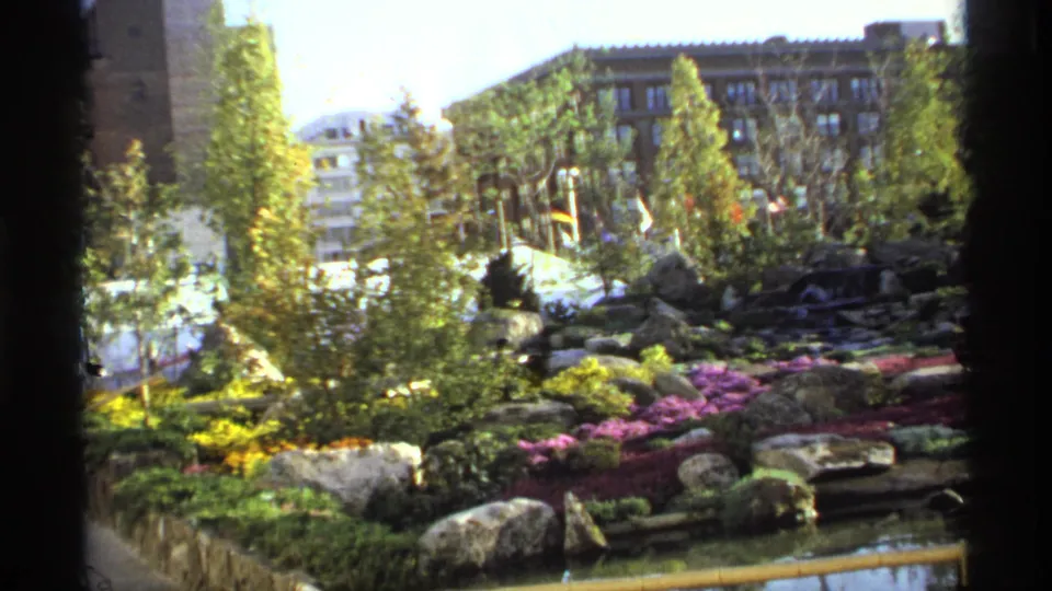 1974 8mm frame of a landscaped garden with water, boulders, bright flower beds, trees, and urban buildings in the background.