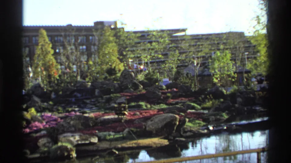 1974 8mm view across a planted garden with rocks, flowers, trees, a small roofed structure, and background buildings.