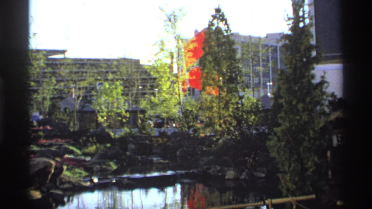 1974 8mm frame of a landscaped garden pond with red flags, evergreen trees, rocks, reflections, and bamboo railing.