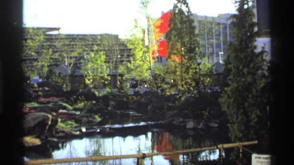 Visitor-shot 8mm view of a garden pond with red flags, rocks, trees, and a modern building facade in the background.