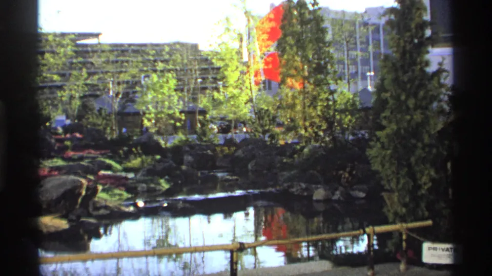 1970s home-movie frame showing a landscaped pond, red flags, evergreen trees, rocks, bamboo railing, and water reflections.