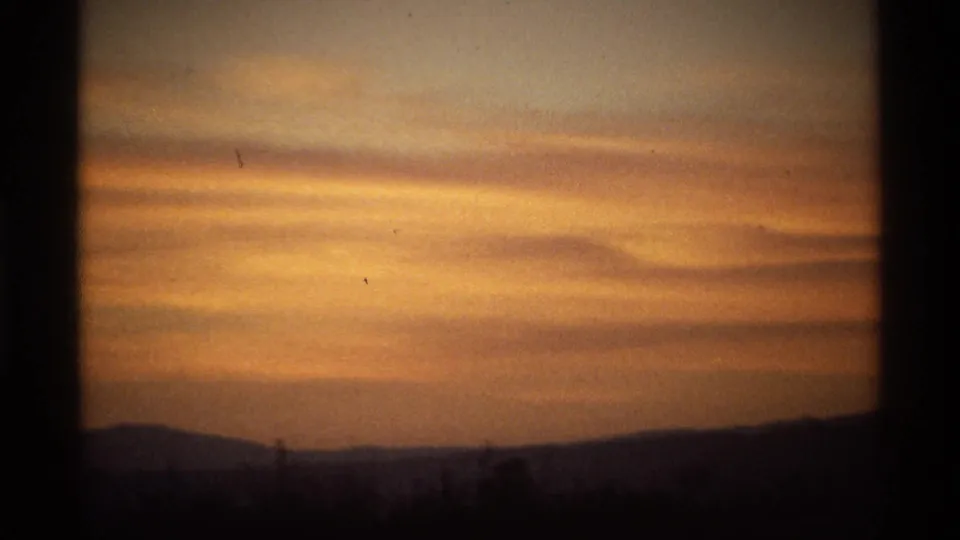 Warm sunset sky with layered orange clouds and a dark silhouetted horizon in archival 8mm footage.