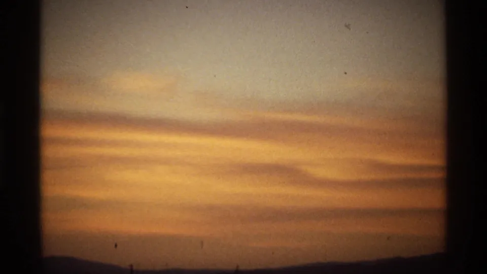 1970s 8mm view of a wide sunset sky with horizontal orange cloud bands and a low dark horizon.