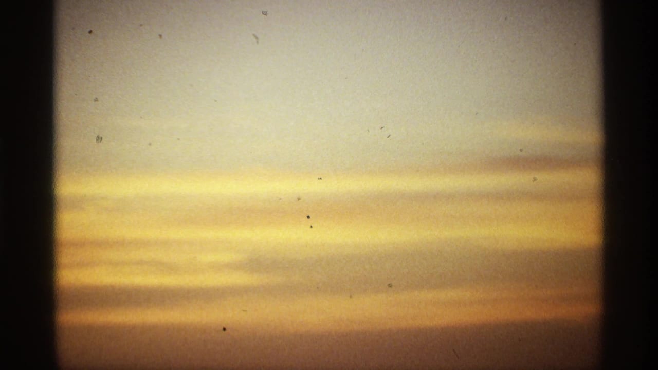 Warm sunrise sky with horizontal cloud bands, film grain, dust specks, and dark frame borders from 8mm home-movie footage.