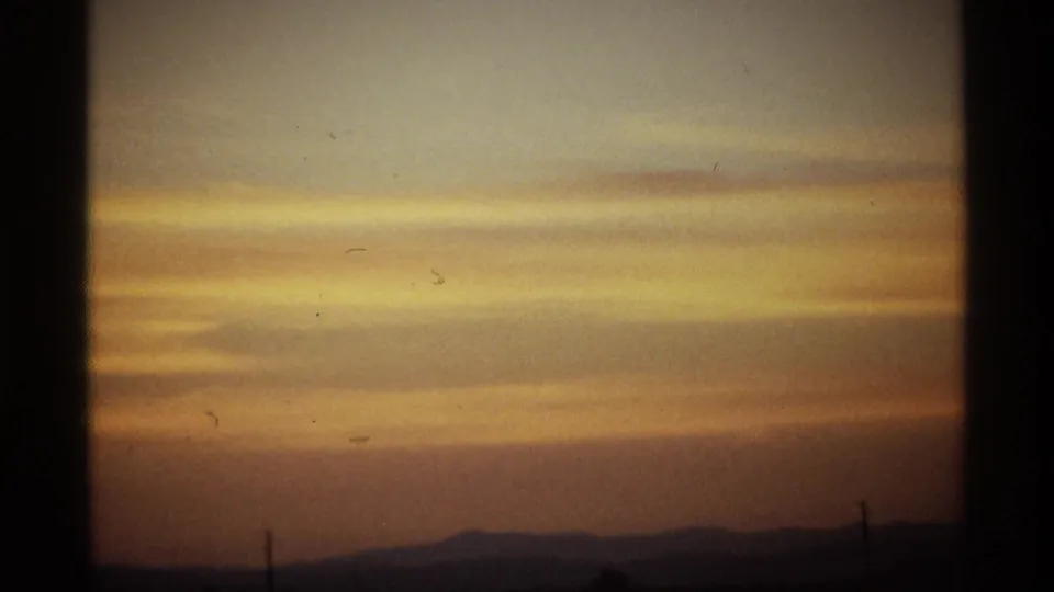 Layered dawn clouds above a dark horizon, with distant hills, a small utility pole silhouette, grain, and vertical film-frame edges.