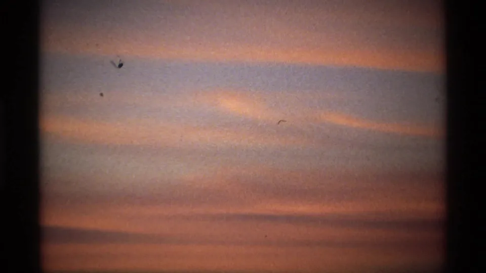 Soft pink-orange clouds stretch across a darkened dawn sky in a grainy 8mm home-movie frame.