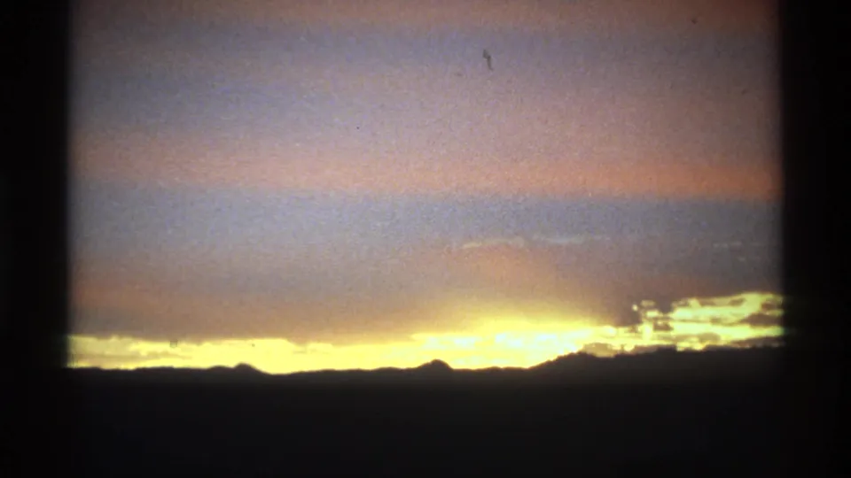 The sun brightens the low horizon under layered pink-gray clouds in a grainy 8mm sunrise shot.