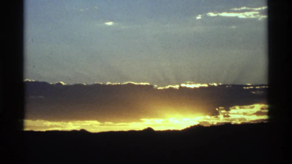 A golden sun glow rises behind a dark horizon as clouds catch light in a soft 8mm frame.