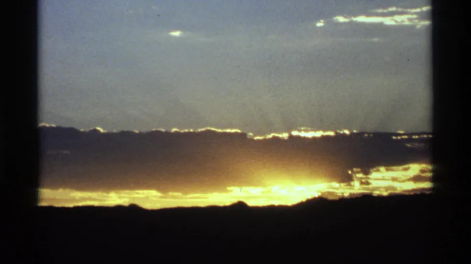 1970s 8mm frame showing a bright low sun glow under a dark cloud band and silhouetted horizon, with visible film edge shadowing.