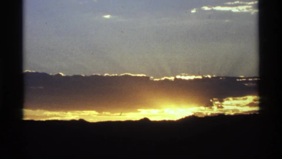 Low sun or sunset light spreads across the horizon beneath layered clouds, with a dark ridge silhouette and 8mm frame-edge shadows.