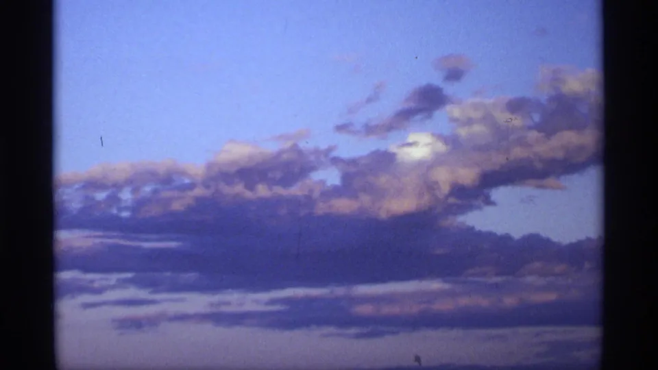 1970s 8mm sky frame with blue upper sky, purple-gold cloud layers, and a dark lower horizon softened by film grain.