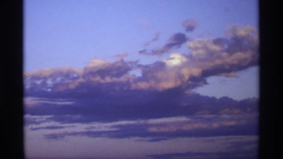 Soft 8mm frame of layered clouds crossing a blue sky, with a low dark horizon and visible color shift from earlier golden light.