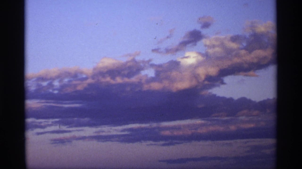 Soft 8mm frame of layered clouds lit by fading evening color, with blue sky, peach highlights, and dark vertical film edges.