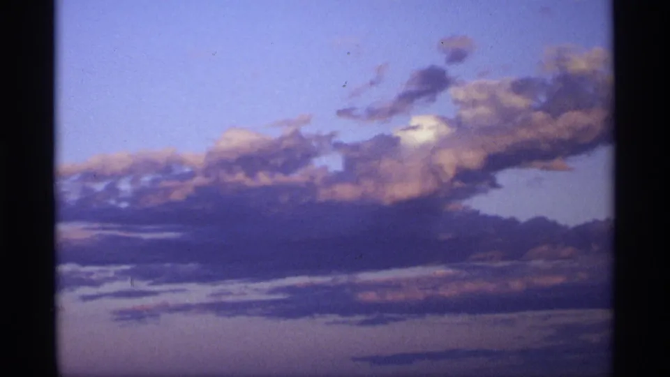 Soft 8mm frame of layered clouds lit by fading evening color, with blue sky, peach highlights, and dark vertical film edges.
