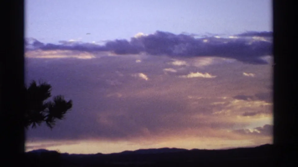 Fading sunset light glows beneath broad clouds above distant dark hills, with a silhouetted tree edge entering from the left.