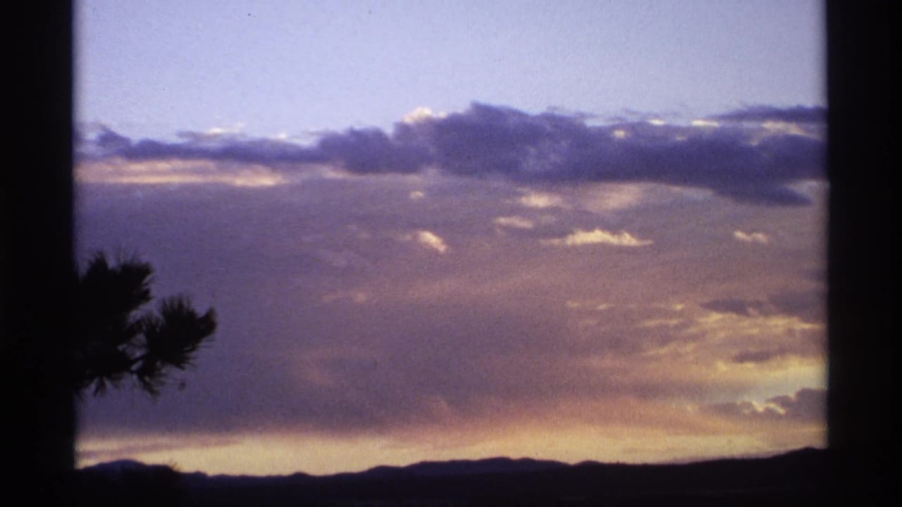 1974 8mm home-movie frame showing clouds and fading light above a dark ridge, with a pine silhouette near the lower left.