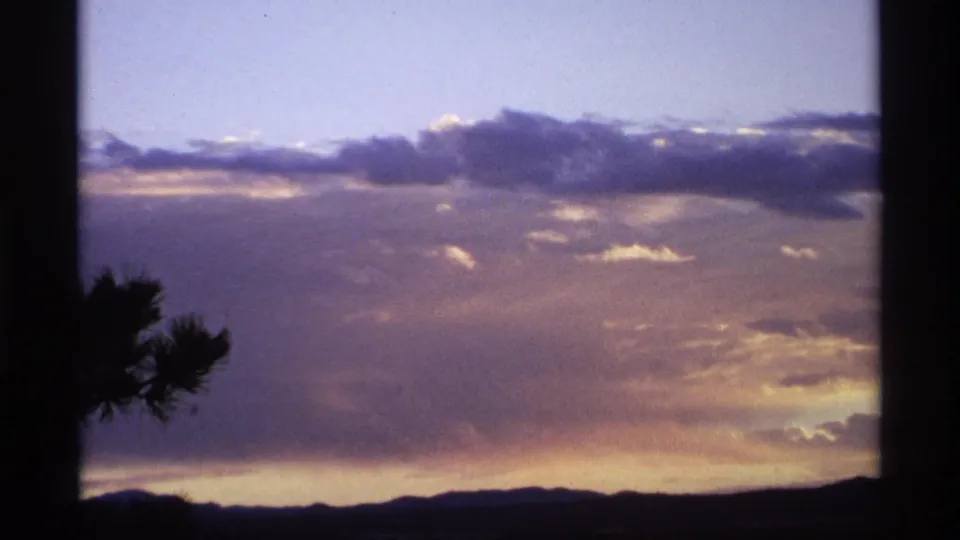 1974 8mm home-movie frame showing clouds and fading light above a dark ridge, with a pine silhouette near the lower left.