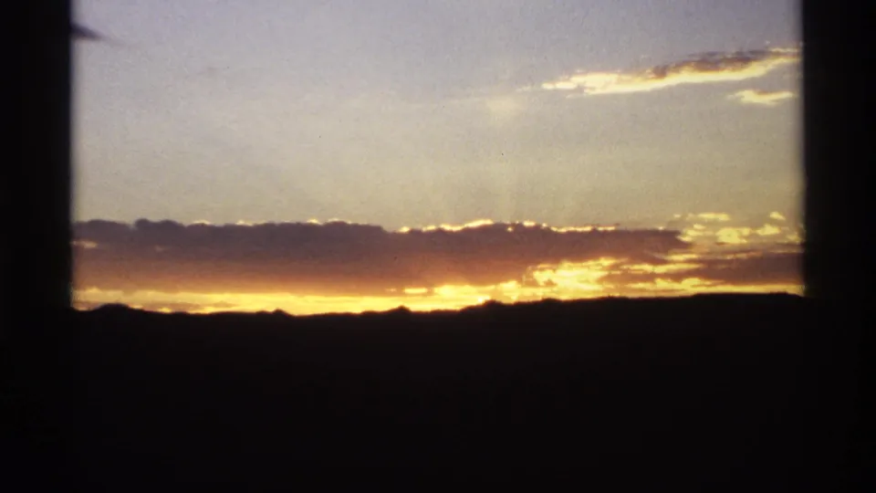 1974 8mm frame with warm orange light along the horizon, cloud bands, and a dark mountain silhouette.