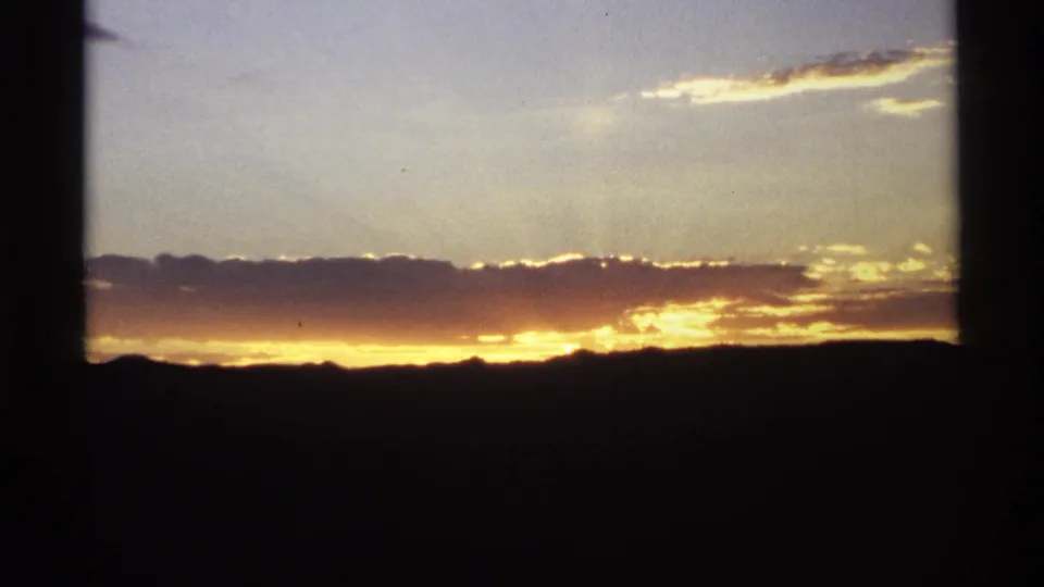 Vintage 8mm frame showing golden sunset light beneath clouds, with a dark ridge line and structural edge at right.