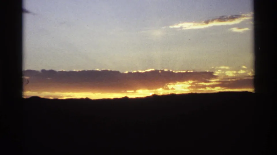 1974 home-movie frame showing a silhouetted ridge beneath glowing clouds and a bright evening horizon.