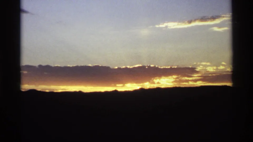 1974 8mm home-movie frame of a red-gold sunset over silhouetted hills, viewed through a dark window frame.