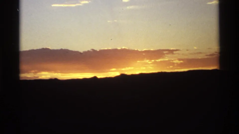 8mm frame showing a brighter orange sunset near the horizon below clouds, with dark mountains silhouetted in the foreground.