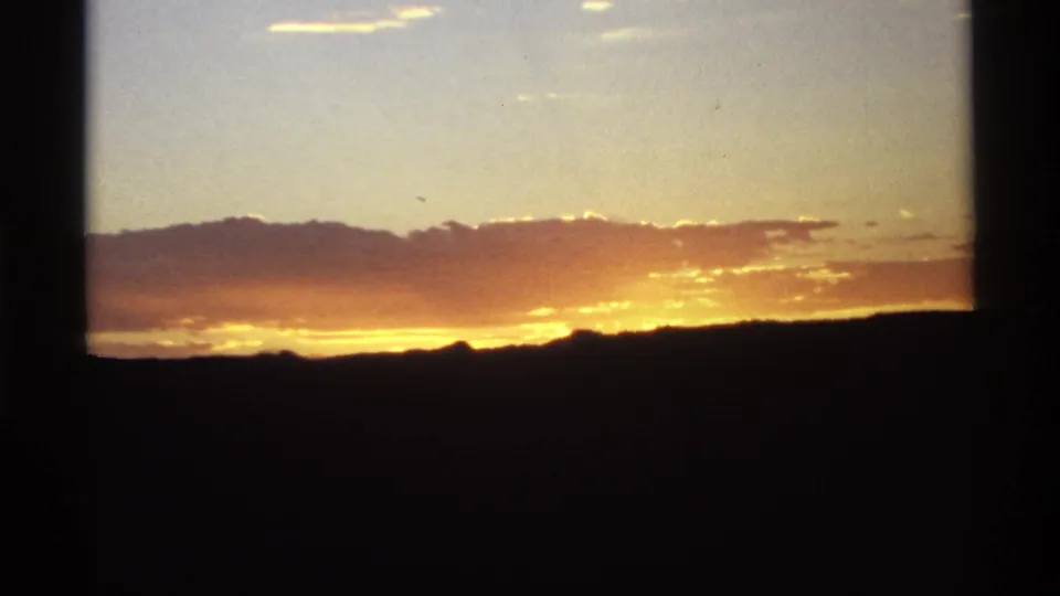 1970s 8mm frame showing a warm sunset glow behind silhouetted mountains and scattered clouds, bordered by dark film edges.