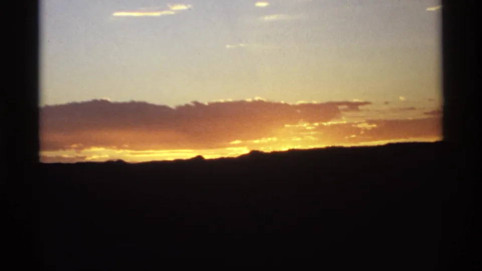 A warm orange sunset spreads across clouds above a dark mountain ridge in archival 8mm home-movie footage.