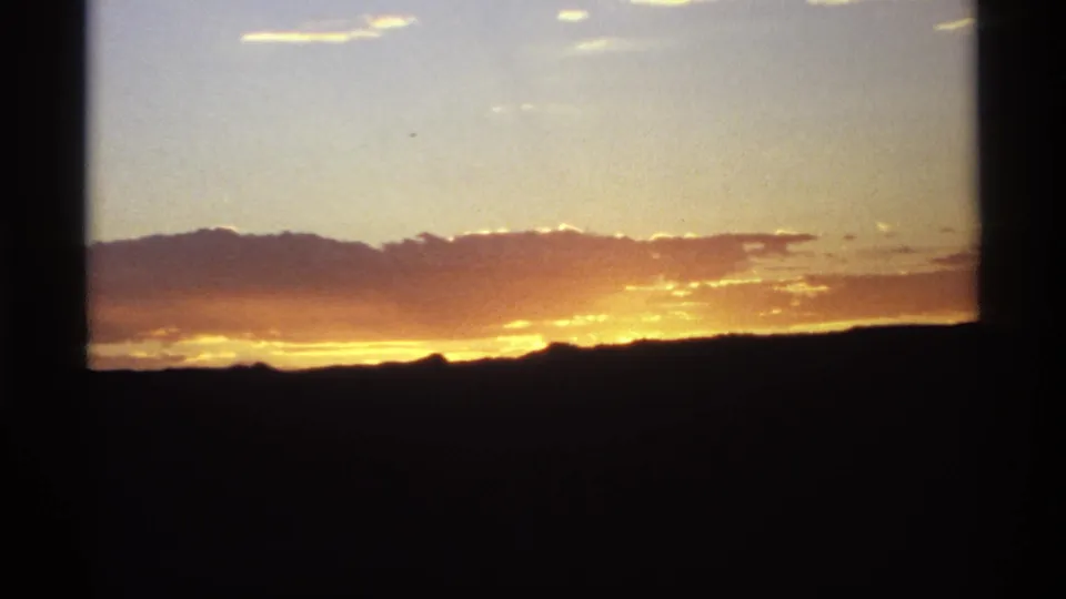 Vintage 8mm frame with a bright golden band of sunset beneath clouds and a nearly black mountain horizon.