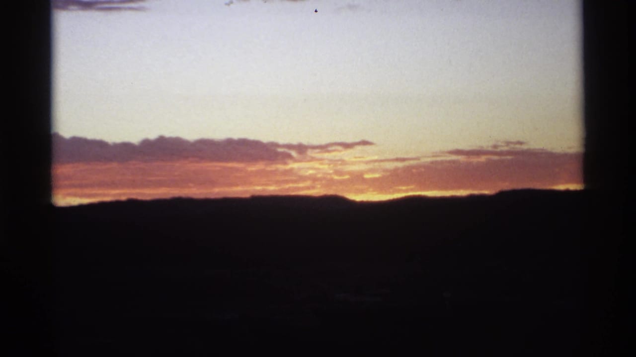 1970s 8mm frame showing a warm sunset behind silhouetted hills, with clouds catching orange light and dark film gate edges visible.