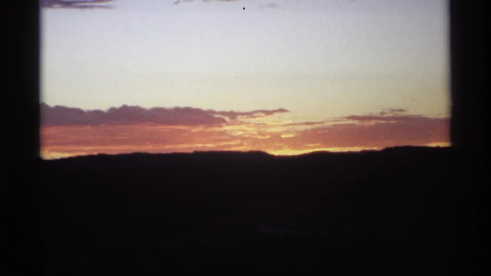 1970s 8mm frame showing a warm sunset behind silhouetted hills, with clouds catching orange light and dark film gate edges visible.