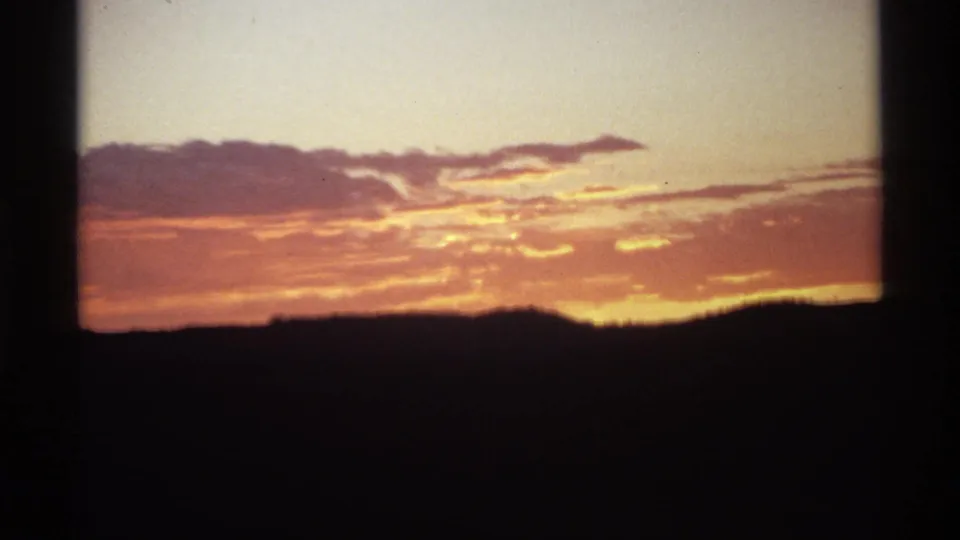 A home-movie sunset view shows orange cloud bands over a dark, uneven horizon with visible film softness and border shadows.