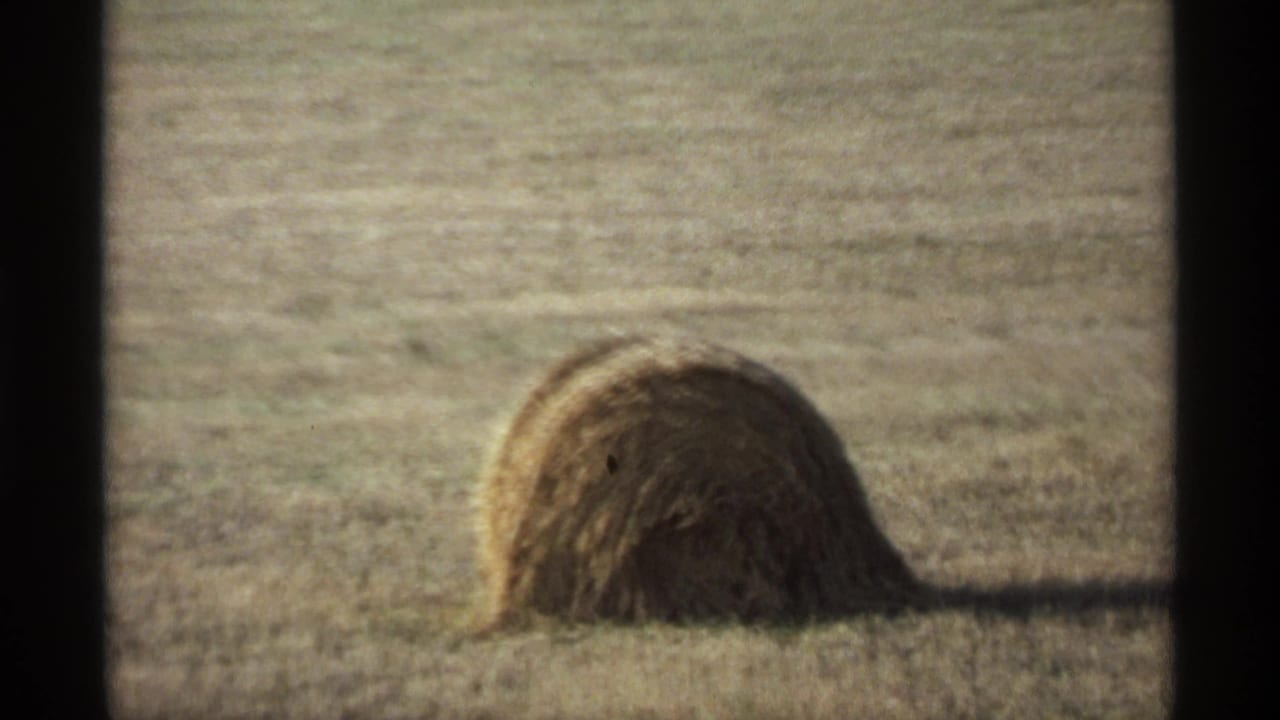 A rounded hay bale sits in a dry field, framed by dark 8mm film borders and soft 1970s home-movie color.