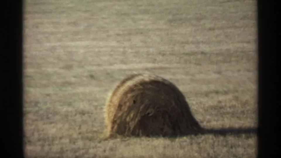 A rounded hay bale sits in a dry field, framed by dark 8mm film borders and soft 1970s home-movie color.