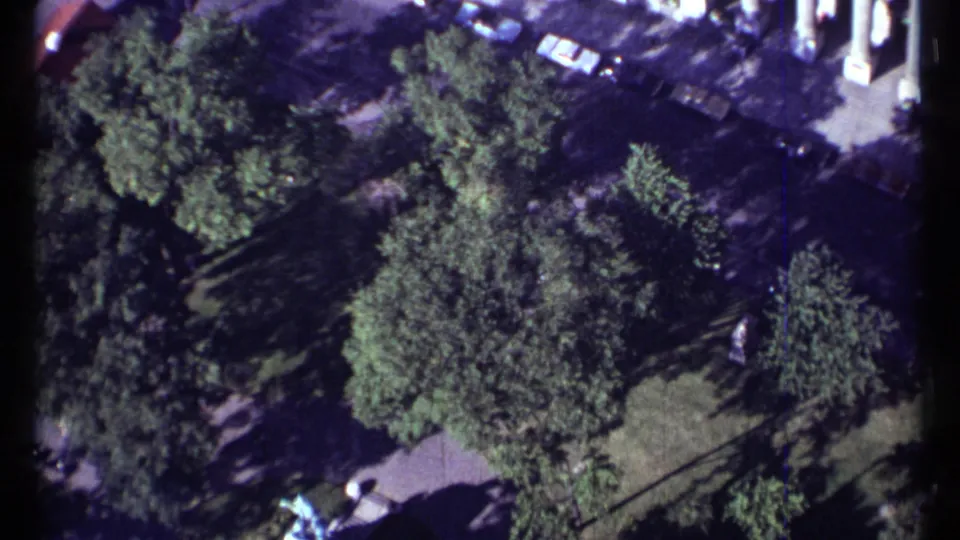 1967 Montreal aerial home-movie frame showing an urban park from above, with trees, lawn, paths, and cars near the street.
