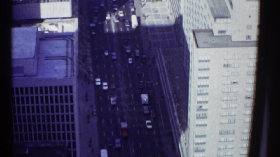 1967 Montreal aerial home-movie frame of a busy street with small cars, tall facades, sidewalks, and gridded building windows.