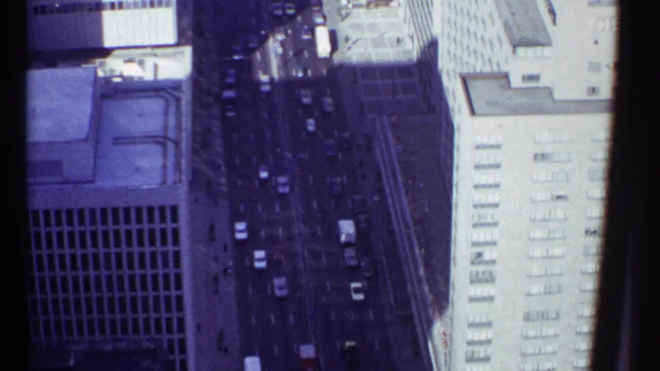 1967 Montreal aerial frame showing traffic on a shadowed city street beside a bright high-rise facade and darker building edge.