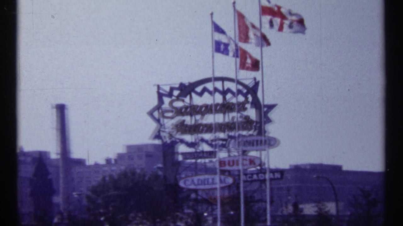Large roadside sign with flags above it and buildings in the background.