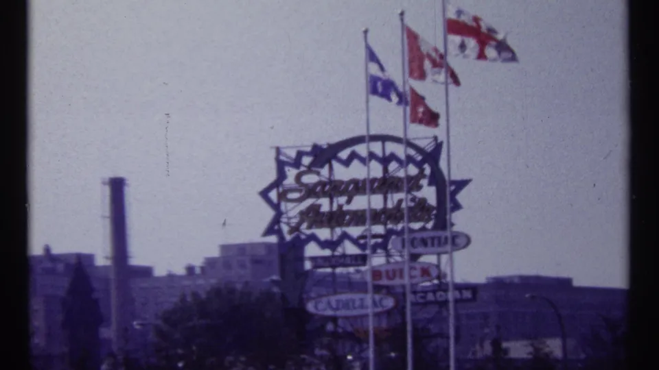 1967 Montreal 8mm frame centered on a large sign, multiple flags, rooftops, and a smokestack in the distance.