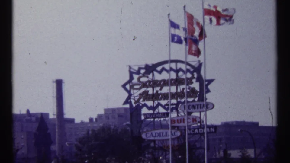 1967 Montreal frame showing a large automobile-related sign, several flags, rooftops, and a distant industrial stack.