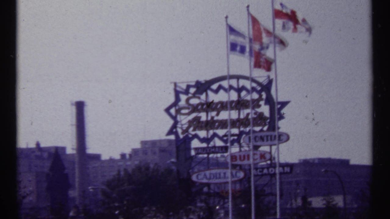 1967 Montreal frame showing a large Saraguay Automobiles sign, multiple flags, and distant buildings in soft 8mm color.